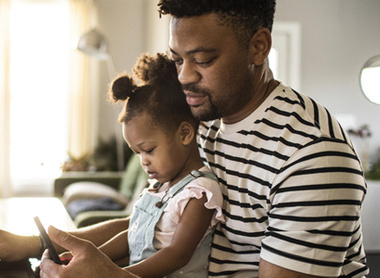 Man with young child on lap, looking at phone and laptop 
